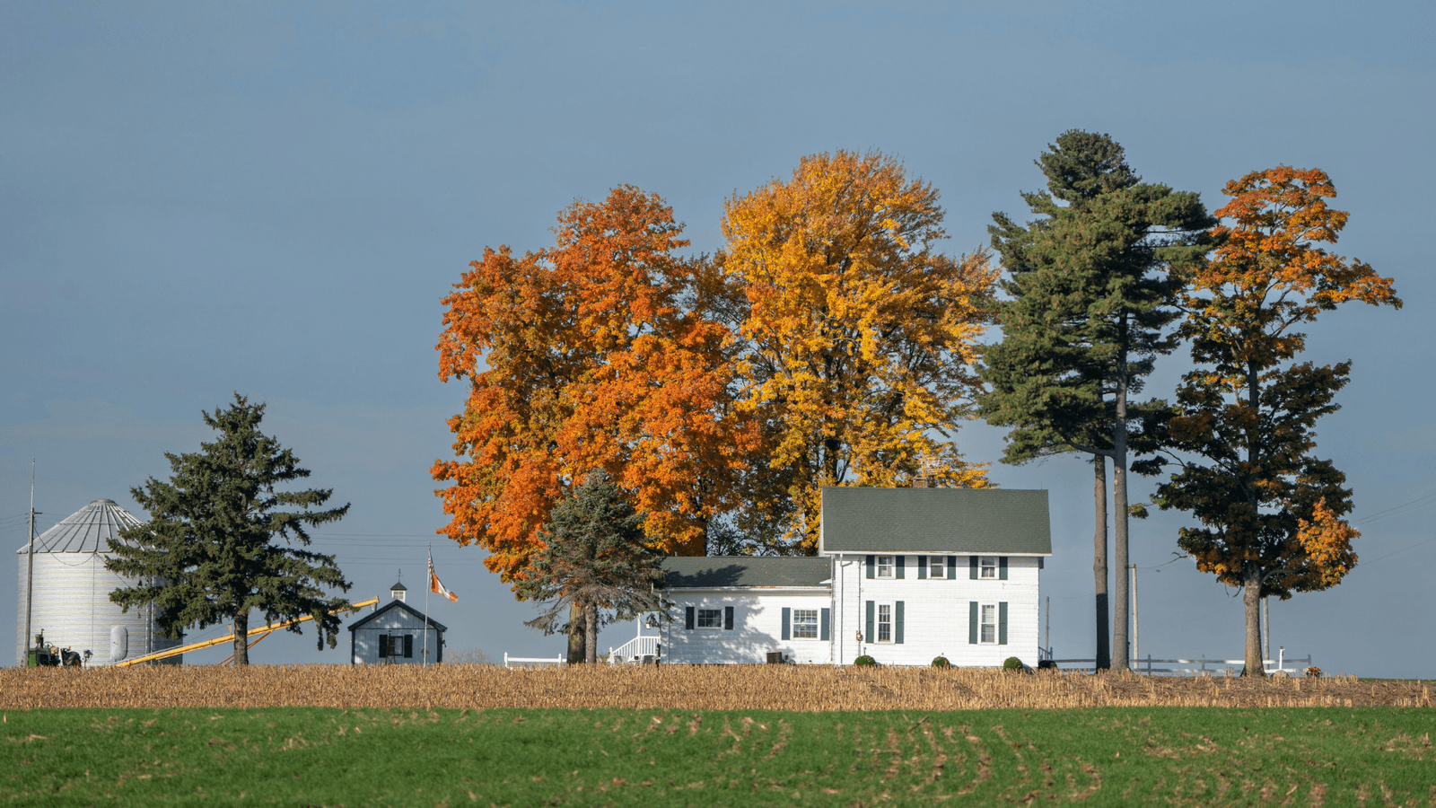 A white Indiana farmhouse beside grain silos with an American flag and autumn trees.
